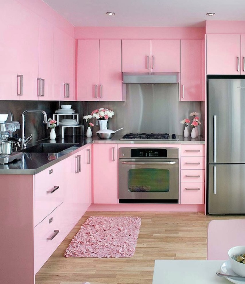 A modern pink kitchen with stylish cabinets, stainless steel appliances, a matching pink rug, wood flooring, and potted flowers brightening the counters.