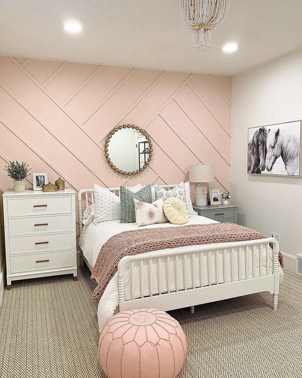 A chic teen girl bedroom featuring a white bed with pink and white bedding, a round mirror on a pink accent wall, a white dresser, and horse artwork displayed above a small nightstand.