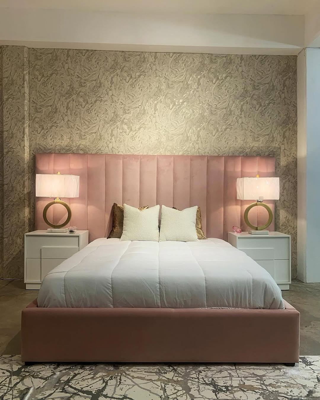 A modern bedroom with a pink upholstered bedroom headboard, white bedding, two white nightstands, gold lamps with round bases, and textured wallpaper behind the bed.