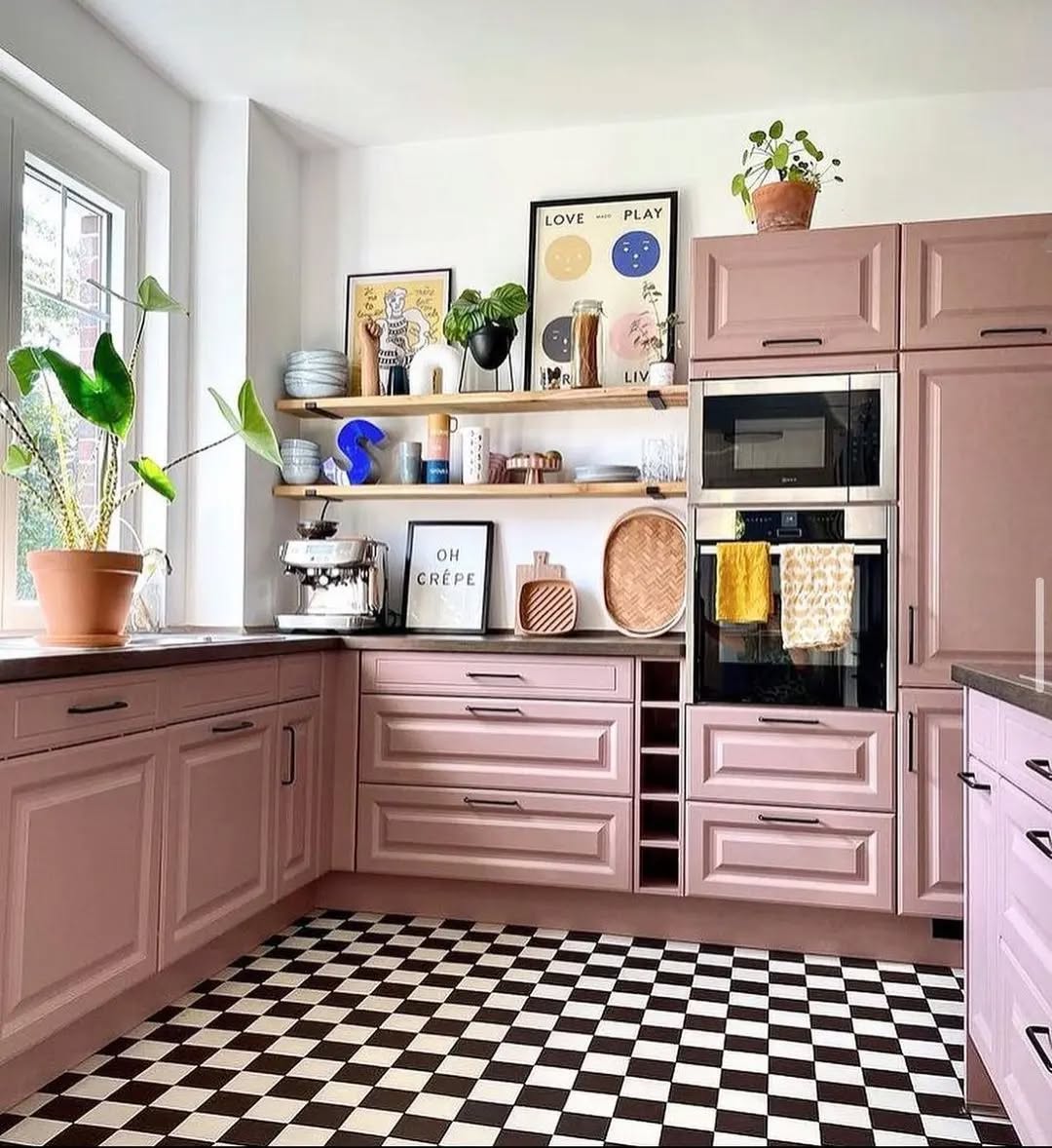 Modern pink kitchen with pastel cabinets, black and white checkered floor, built-in appliances, potted plants, open shelving, and decorative artwork on the wall.