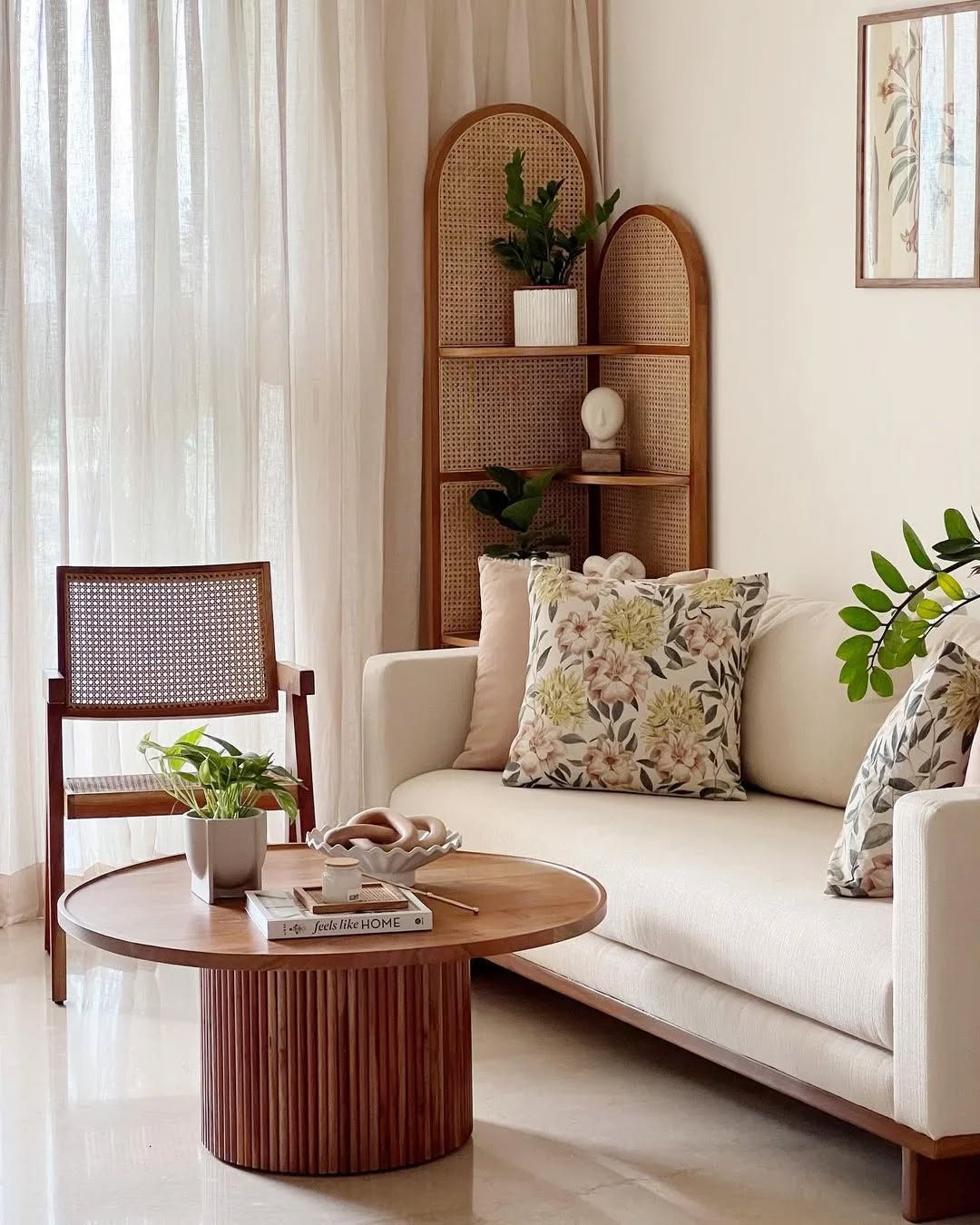 A modern living room with a white sofa, floral cushions, a round wooden coffee table, potted plants, and a cane chair offers stylish interior design inspiration and chic corner décor against a white wall and sheer curtains.