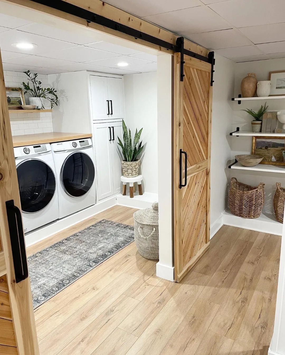 A modern cottage laundry room with a front-load washer and dryer, wooden sliding barn door, potted plants, shelves with baskets, and light wood flooring.