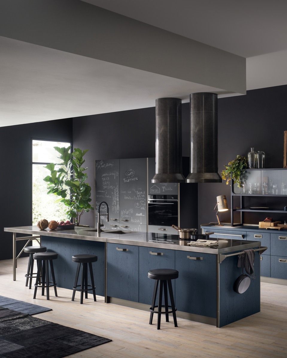 A rustic industrial kitchen features a stone wall, copper light fixtures, a copper range hood, wooden cabinets, and a central island with a concrete countertop and exposed beams.