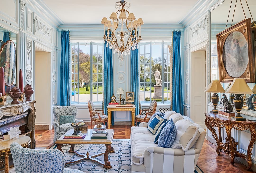 A formal french country living room with elegant blue accents, ornate furniture, a chandelier, large windows with blue curtains, and views of a garden with a statue.