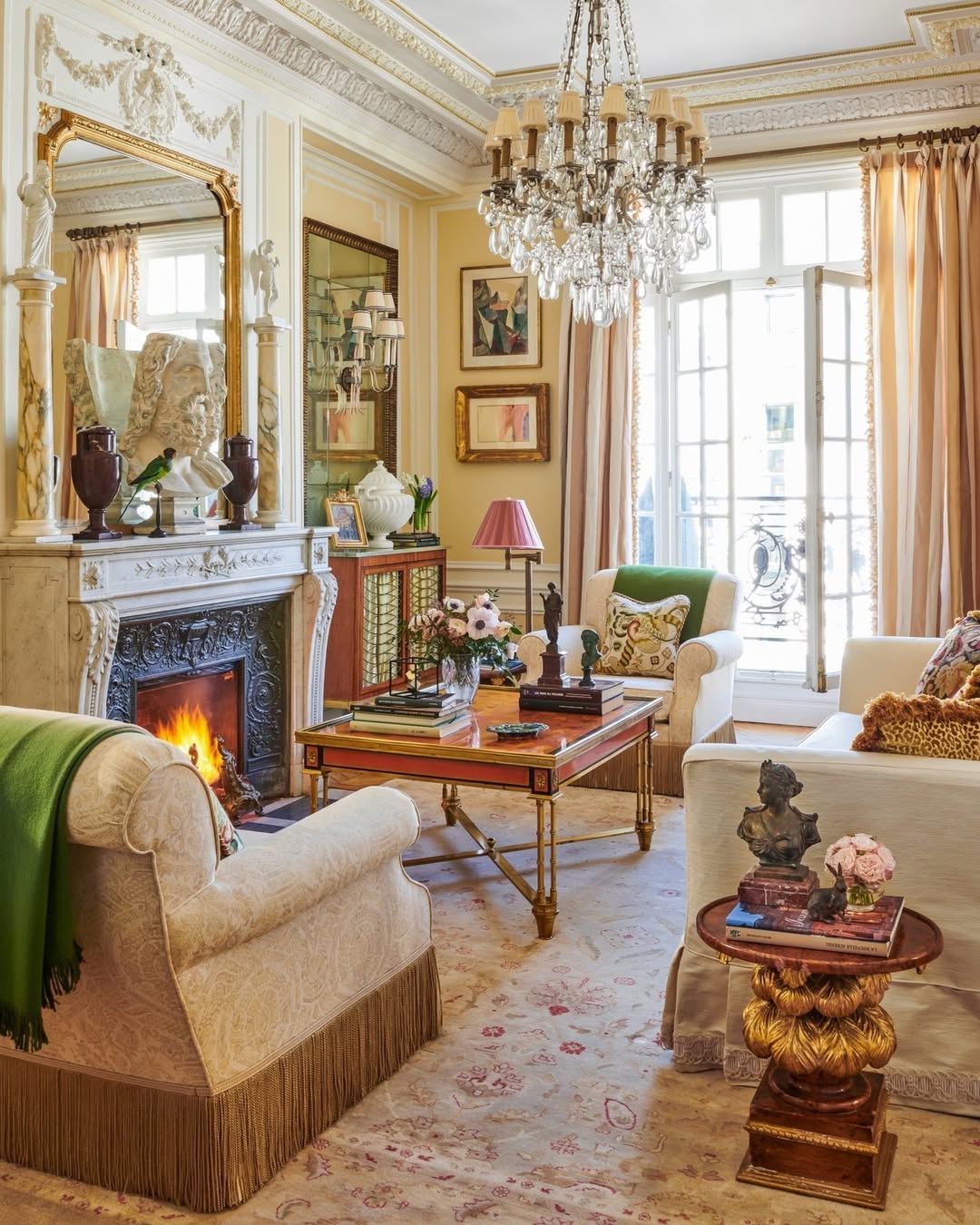 Elegant french country living room with ornate molding, fireplace, chandelier, and antique furniture; sunlight streams through French doors onto a richly decorated space with artwork and decor accents.