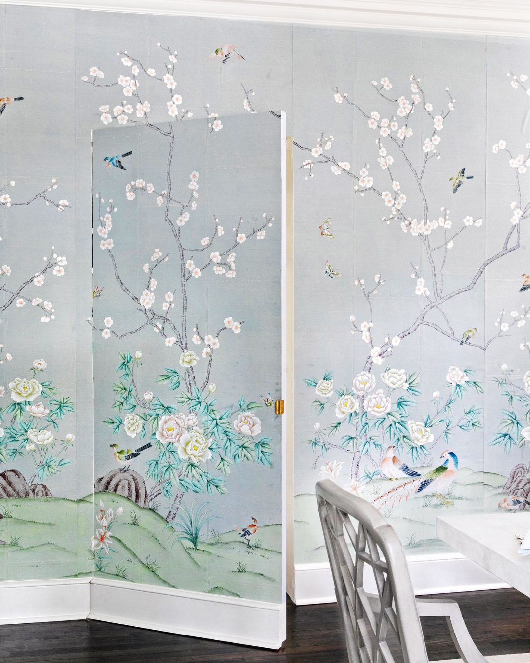 A hidden door blends into a wall covered with pale blue floral wallpaper featuring white blossoms, green foliage, birds, and a partially visible white chair and table—perfect inspiration for stylish homeowners seeking creative hidden door ideas.