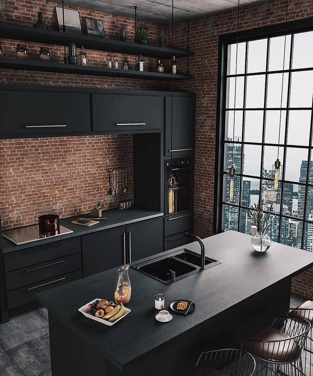Modern industrial kitchen with black cabinets and island, brick walls, large industrial windows offering a city skyline view, and a tray of food and drinks on the counter.