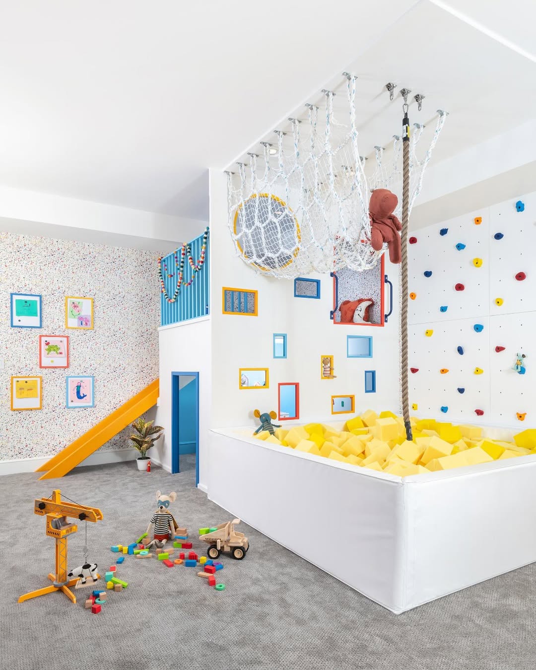 Under stair playroom featuring a yellow slide, climbing wall, rope ladder, net, stuffed toys, foam pit, wooden toys, and colorful framed pictures on patterned walls—perfect for endless children’s adventures.