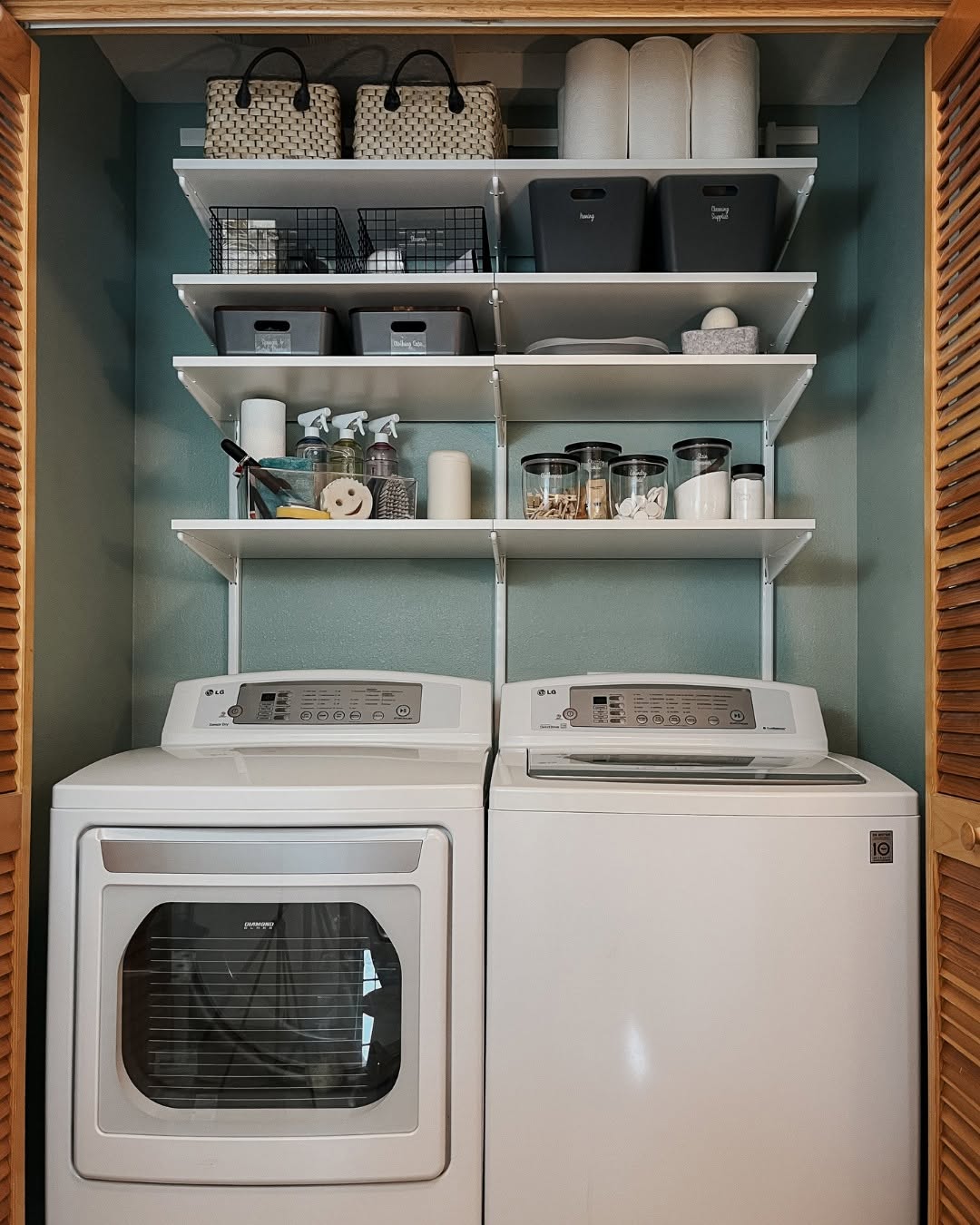 A laundry area with a washing machine and dryer beneath four organized Laundry Room Shelves holding baskets, bins, cleaning supplies, and jars against a green wall.