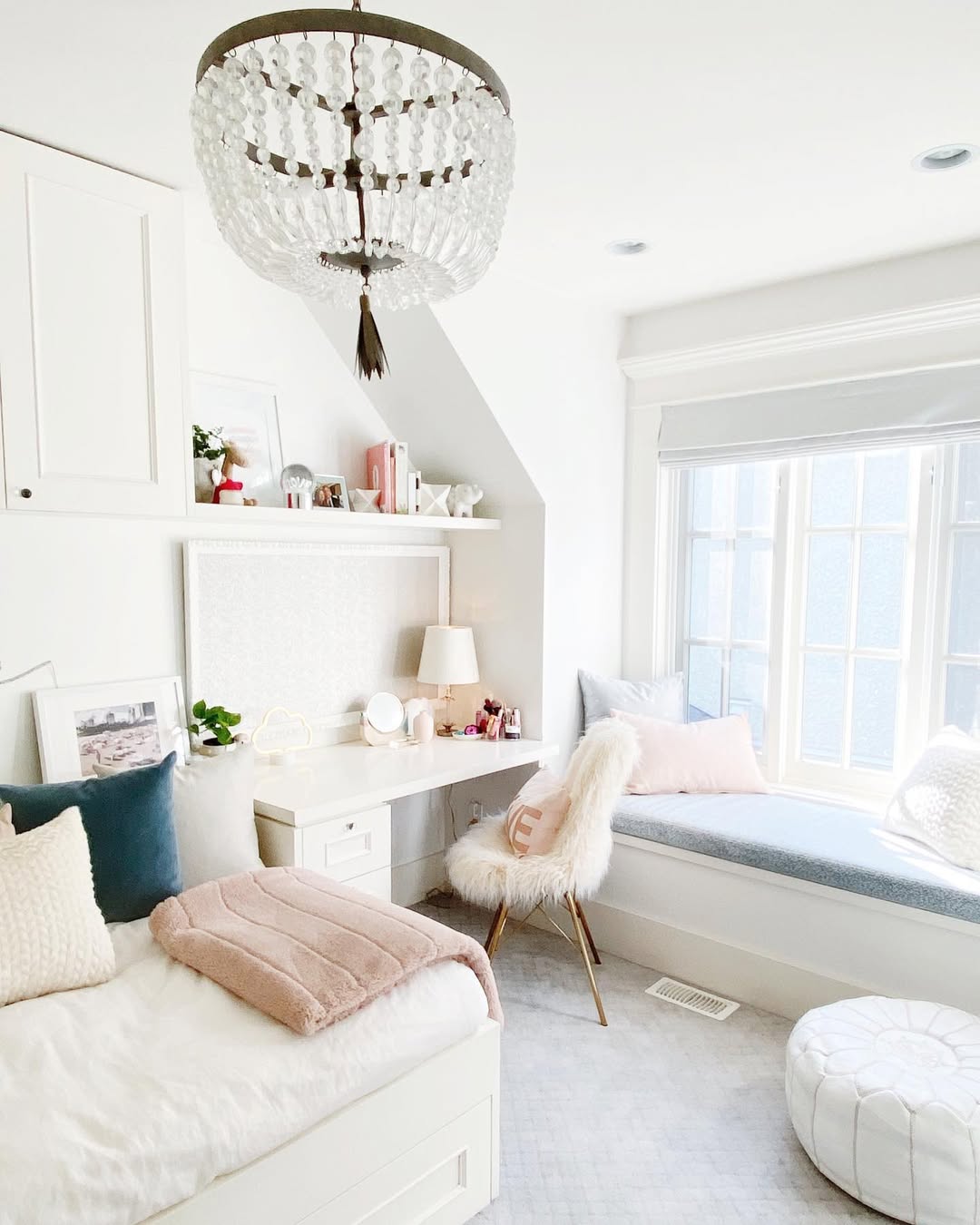 A bright, white teen girl bedroom with a bed, desk, fur-covered chair, window seat, and decorative chandelier. Soft pastel pillows and throws add warmth to the space.