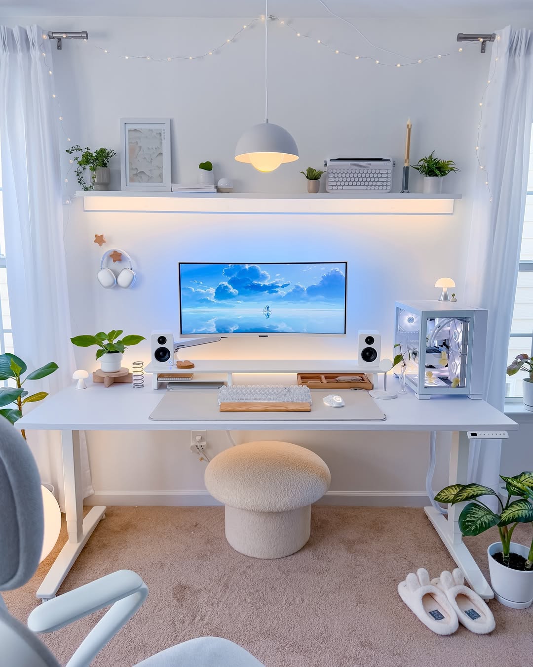 Minimalist white computer desk setup for a small home office, featuring a large monitor, mechanical keyboard, plants, decor items, and LED lighting; cozy stool and slippers rest on a beige carpet.