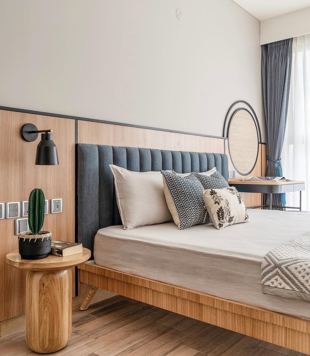 Modern bedroom with a wooden bed frame, stylish bedroom headboard in grey upholstery, patterned pillows, a round wooden side table, cactus plant, wall lamp, and large window with curtains.