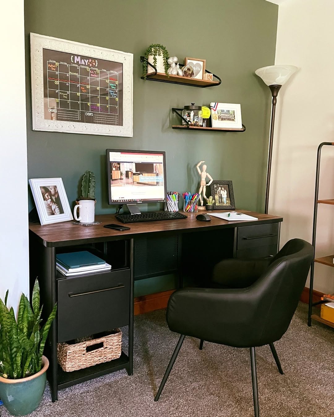 A modern small home office with a desk, computer, black chair, lamp, wall calendar, shelves with decor, framed photos, and a potted plant on a gray carpeted floor.