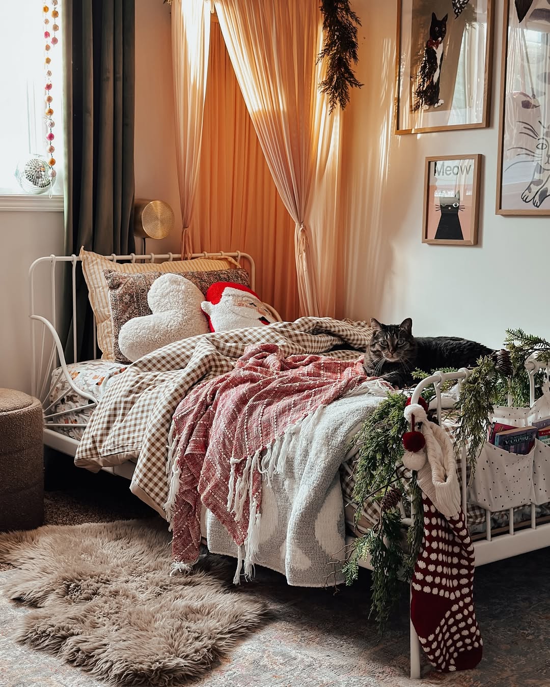 A cozy teen girl bedroom with a neatly made bed, layered blankets, pillows, a cat lounging on the bed, and Christmas decorations hanging from the bedframe and wall.