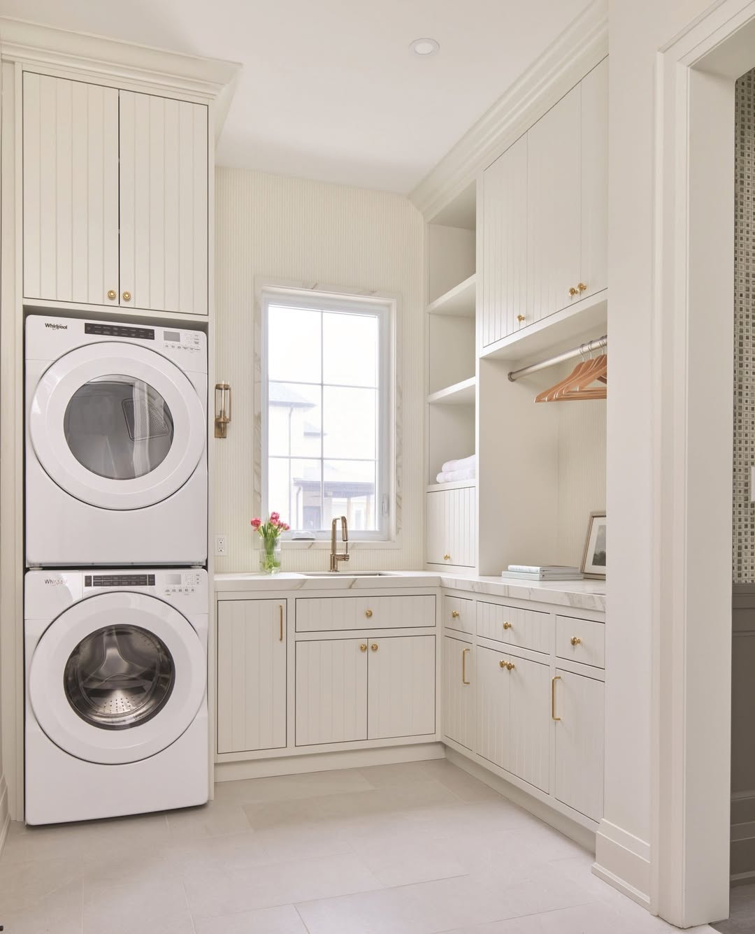 A modern cottage laundry room with a stacked washer and dryer, white cabinetry, gold hardware, a sink under a window, and open shelves with hangers and towels.