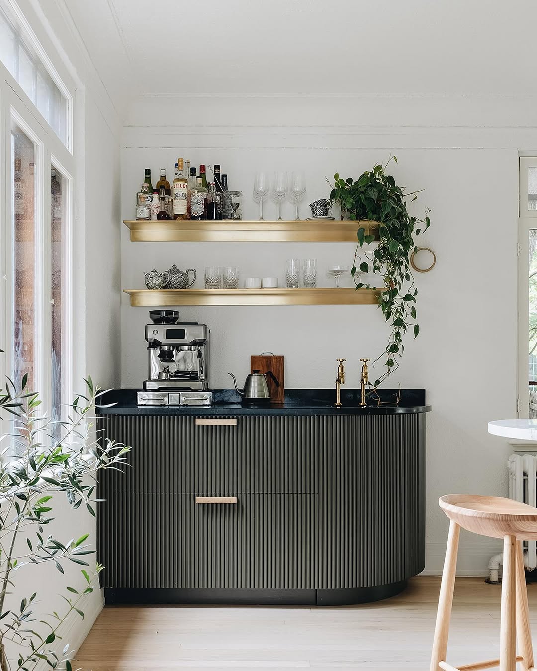 Modern home coffee bar with two gold shelves holding bottles, glassware, and a plant above a black countertop with a coffee machine and kettle, set against a white wall—perfect for those seeking stylish home coffee bar ideas.