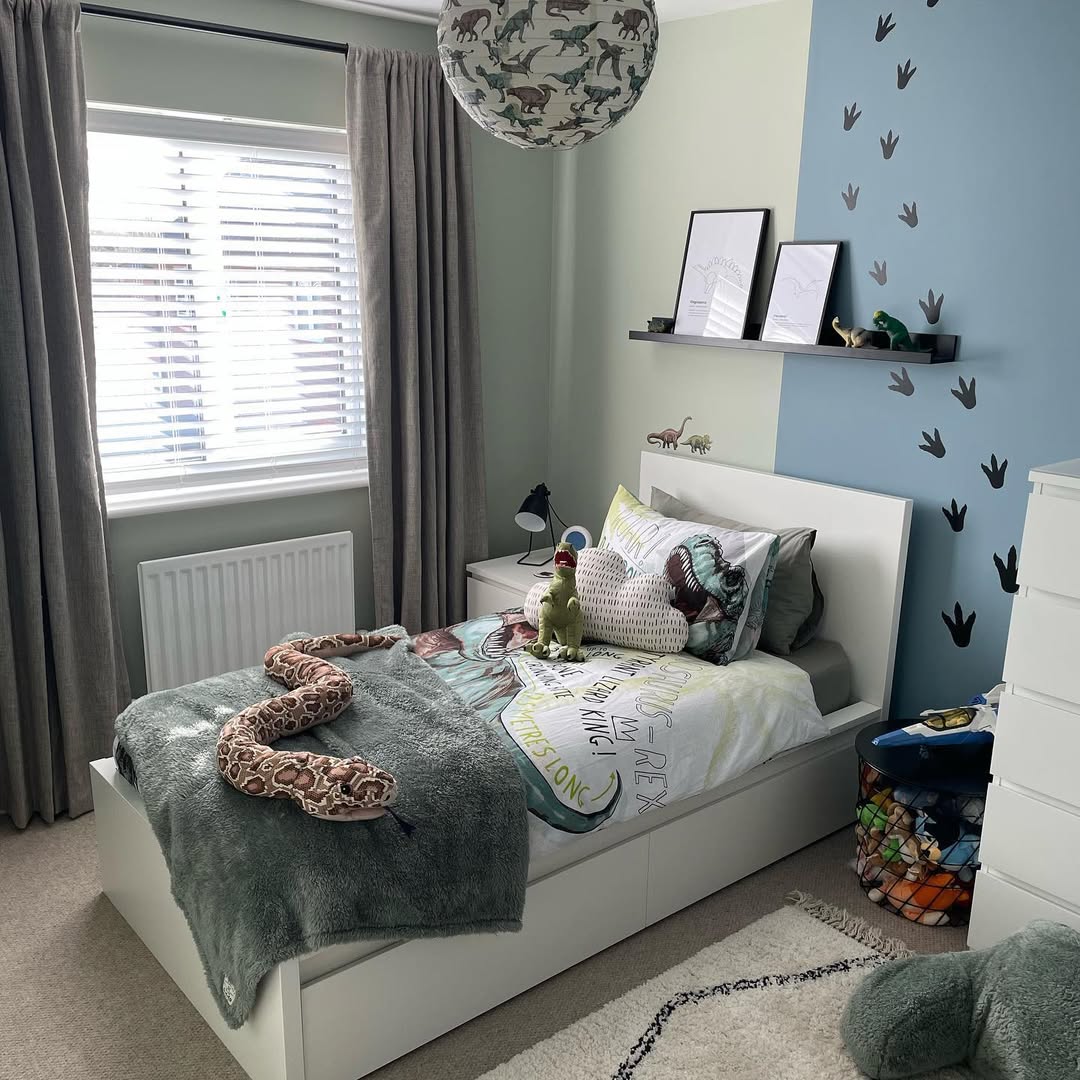 A child's dinosaur bedroom comes alive with themed decor, showcasing a bed draped in dinosaur bedding and toys. The walls proudly display vibrant dinosaur prints, while shelves hold framed pictures. Light gently filters through the window blinds, completing this prehistoric paradise.