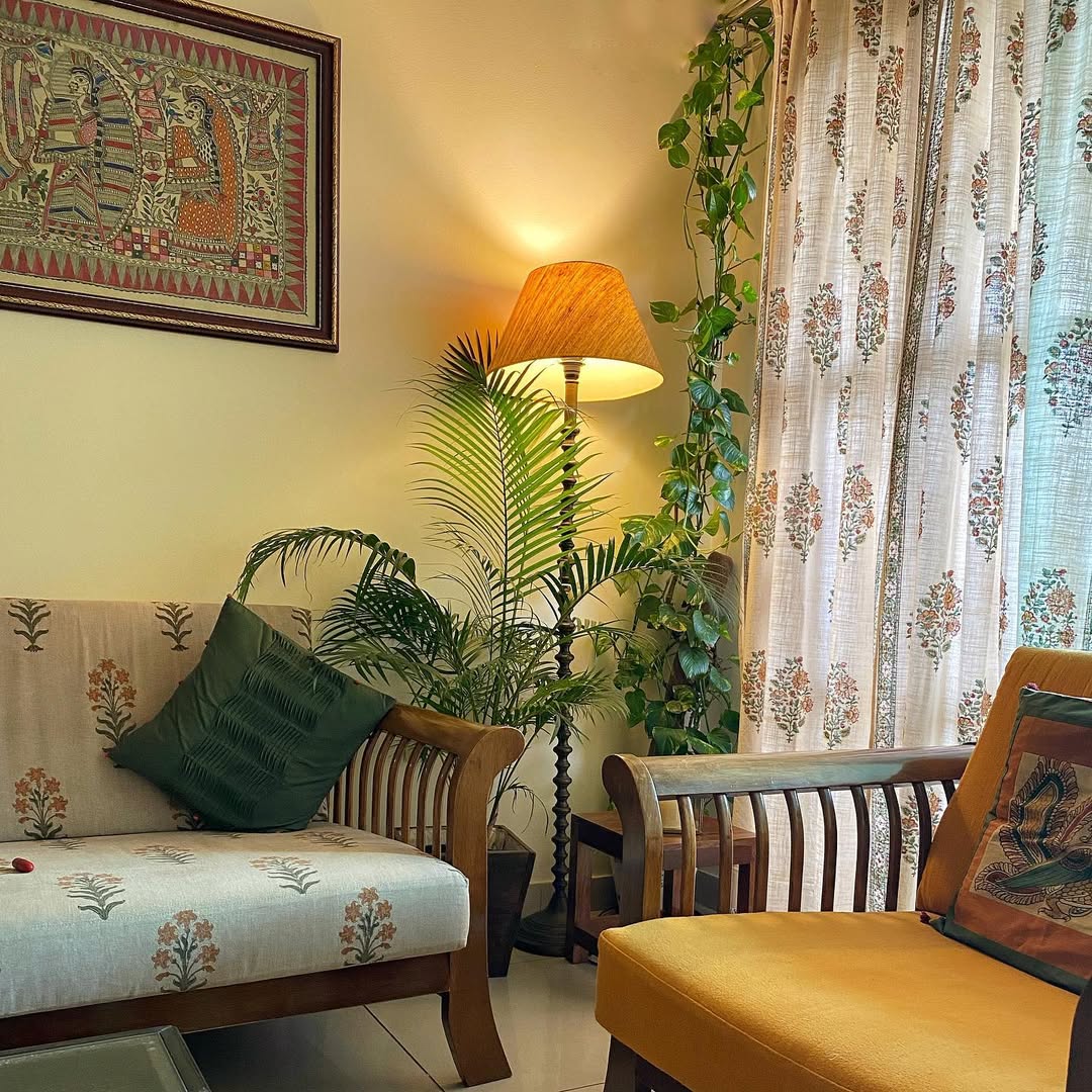 A cozy living room with wooden furniture, patterned cushions, indoor plants, and a lit floor lamp offers inspiring corner decor. Sheer floral curtains by the window complete this inviting home decoration.