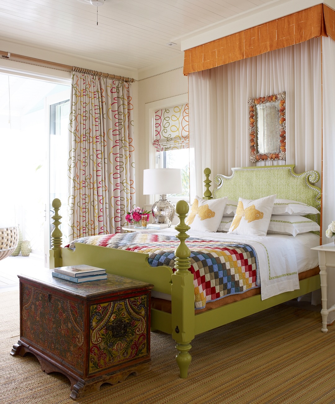 A brightly decorated bedroom with a green four-poster bed featuring an intricate headboard, a patchwork quilt, ornate chest, and patterned curtains beside a large window with a hanging chair outside.