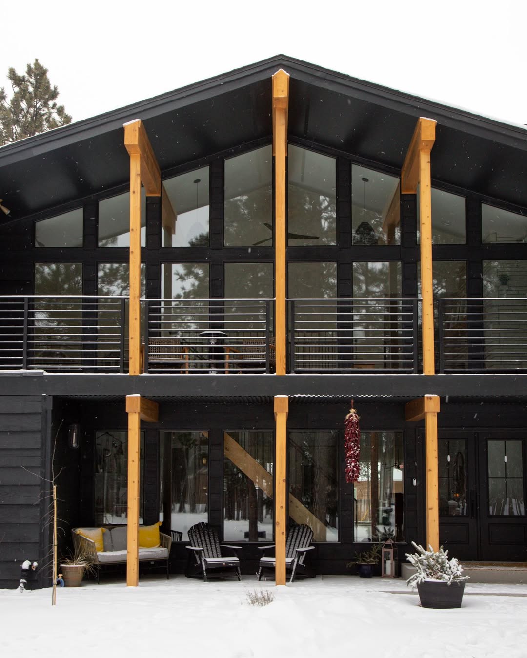 Modern two-story black and cedar house exterior with large glass windows and wooden beams, surrounded by snow, featuring stylish outdoor seating on the patio and balcony—an inspiring option for those seeking unique house exterior ideas.