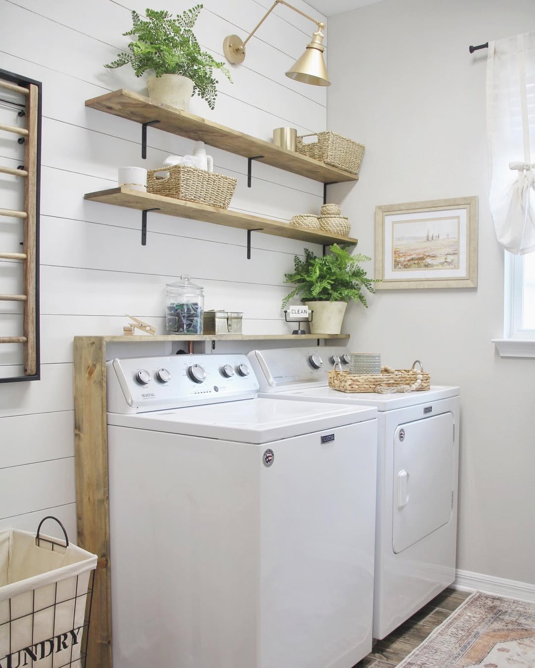 A bright laundry room with a washer and dryer, wooden laundry room shelves holding baskets and plants, a framed picture on the wall, and a laundry basket on the floor.