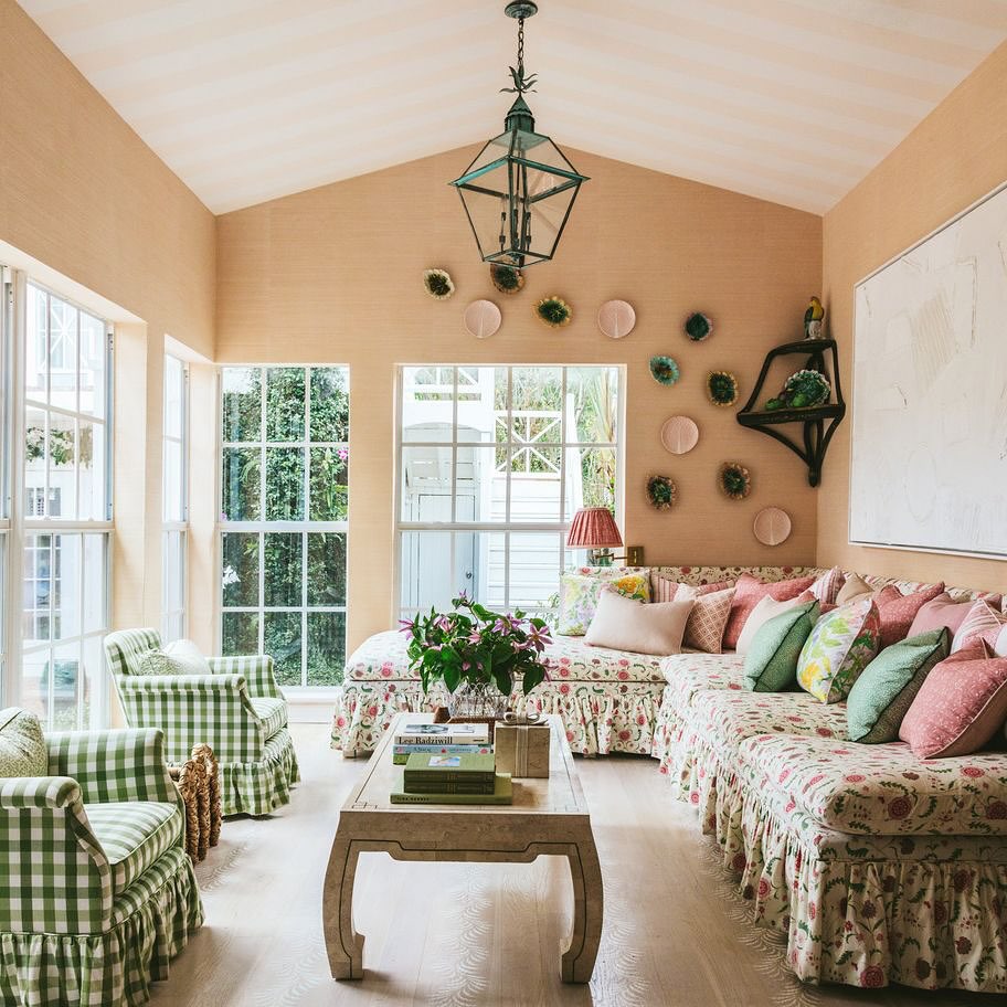 A sunlit french country living room with floral and gingham furniture, a large sectional sofa, a wooden coffee table, and decorative plates on the wall.