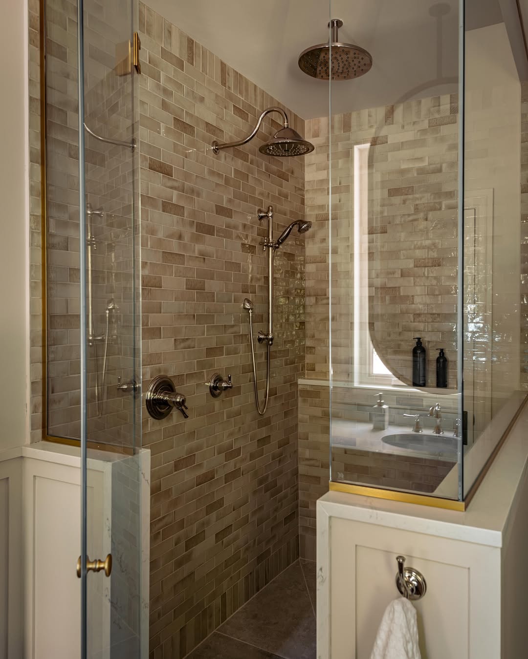 Modern glass-enclosed, no door shower with beige subway tile, rainfall and handheld showerheads, wall niche, and metal fixtures; two black bottles on a ledge near a mirror offer stylish walk-in shower ideas.