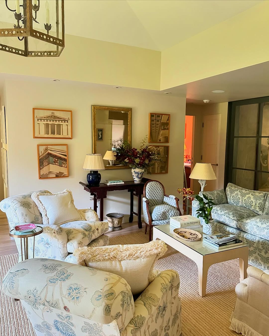 A french country living room with patterned armchairs, a floral sofa, a coffee table, lamps, framed art on the wall, and a console table topped with a flower arrangement.