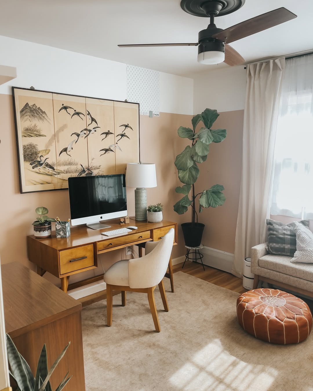 A home office with a wooden desk, computer, chair, potted plant, lamp, large wall art, pouf, and a window with sheer curtains letting in natural light—perfect for those seeking minimalist office ideas.