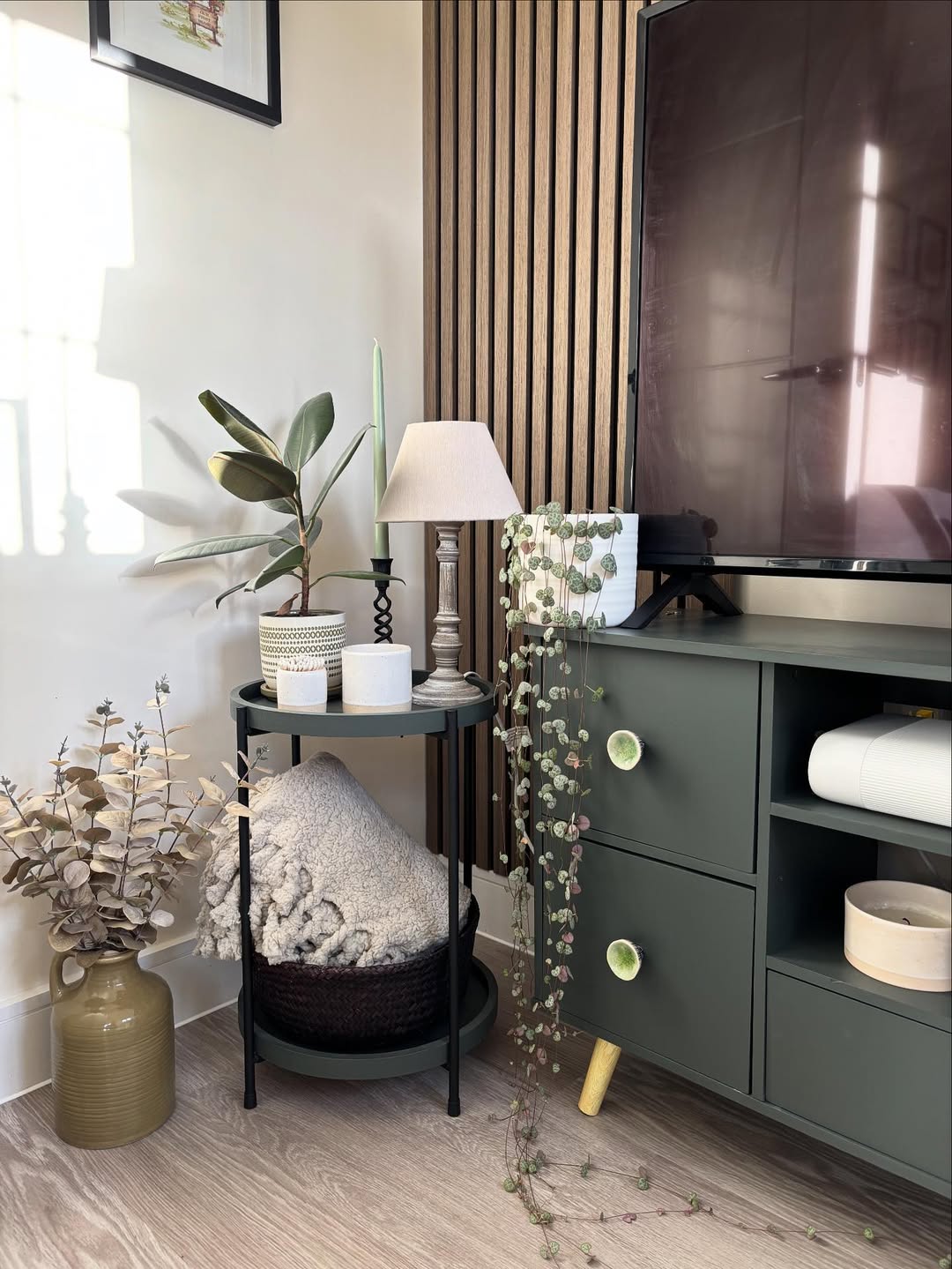 A living room corner with a green TV stand, round side table holding a lamp and candles, potted plants, a rolled blanket, and decorative vases with dried leaves—perfect inspiration for stylish corner decoration ideas in home decor.