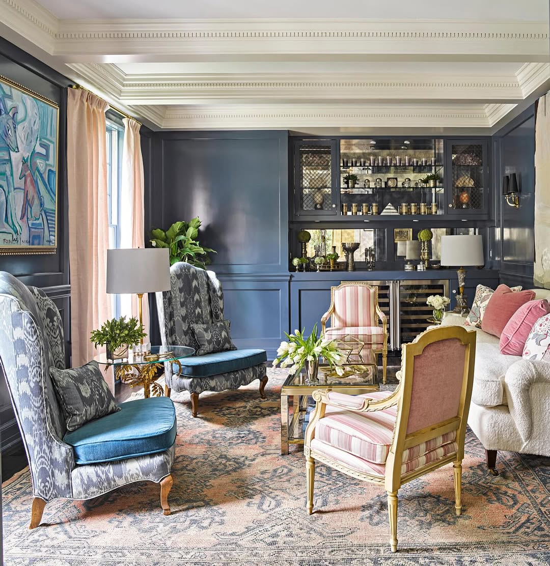 A traditional French country living room with blue paneled walls, patterned armchairs, a glass coffee table, built-in shelves, and large windows with curtains.