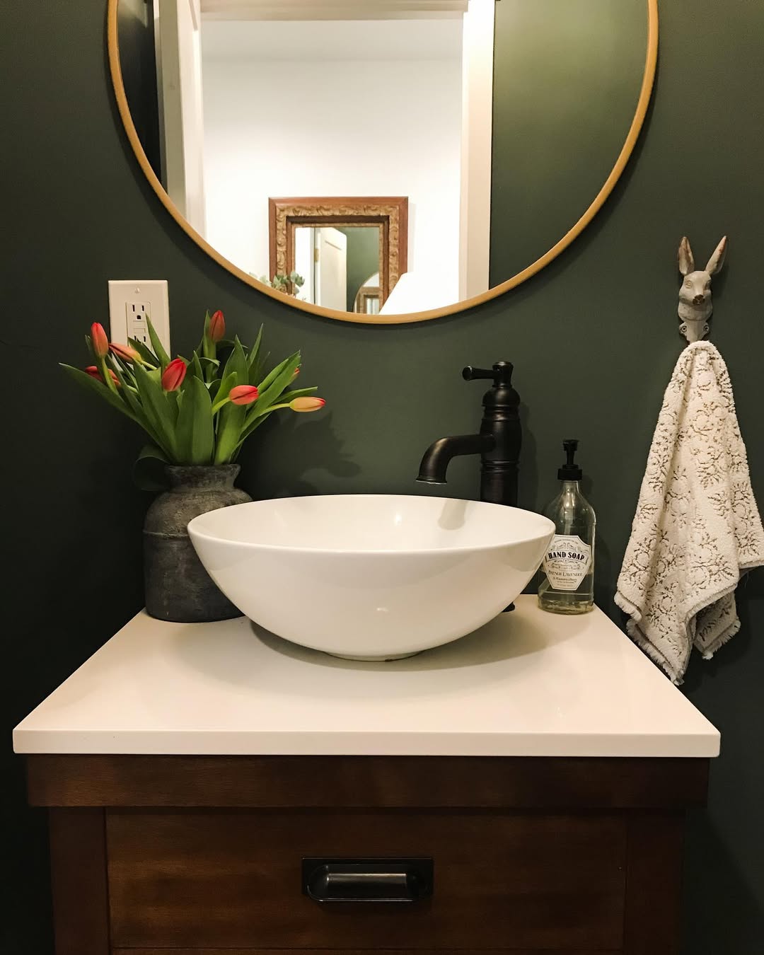 A modern bathroom vanity, perfect for small bathroom decor, features a white vessel sink, black faucet, vase of red tulips, hand soap, round mirror, and a towel on a rabbit head wall hook.