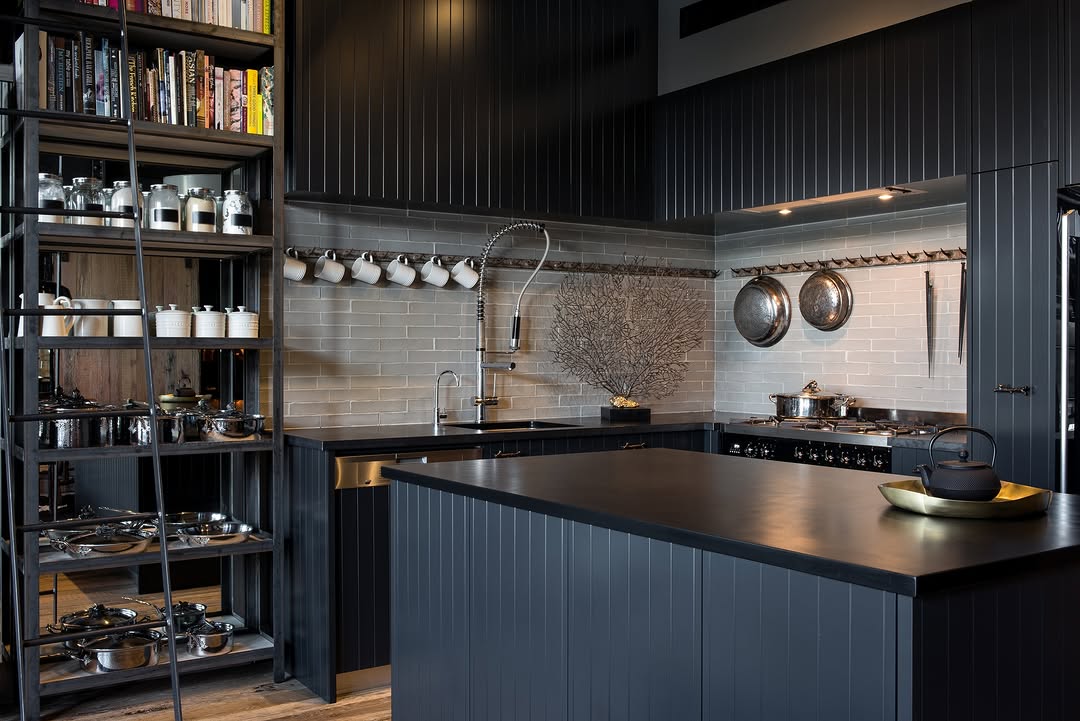Modern industrial kitchen with dark cabinetry, open shelving displaying cookware, a tile backsplash, and a large central island featuring a black teapot on a tray.
