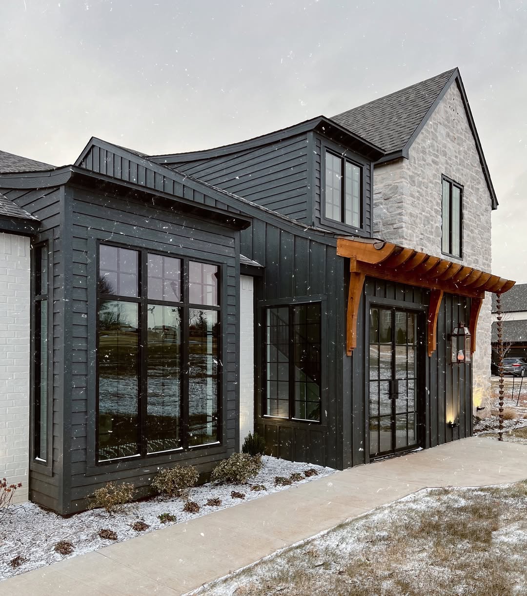 Modern two-story house with black house exterior, stone accents, large windows, and a wooden pergola over the entrance; light snow on the ground and sidewalk.