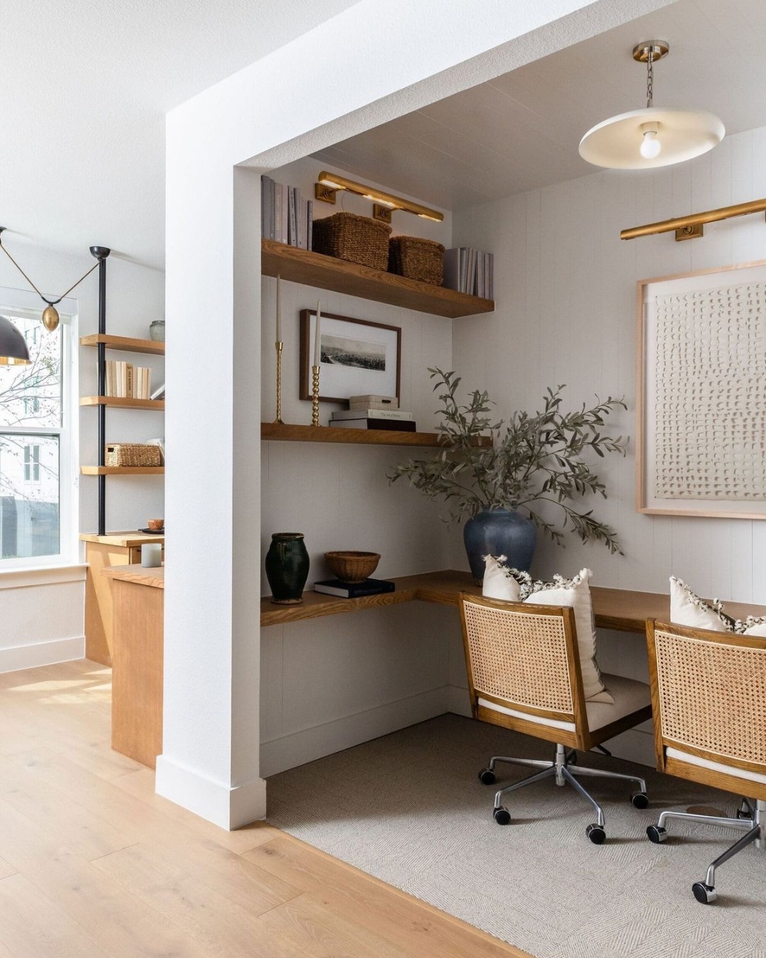 A modern small home office with two rattan chairs, a wooden desk, floating shelves with decor, framed art, a blue vase with greenery, and neutral-toned furnishings.