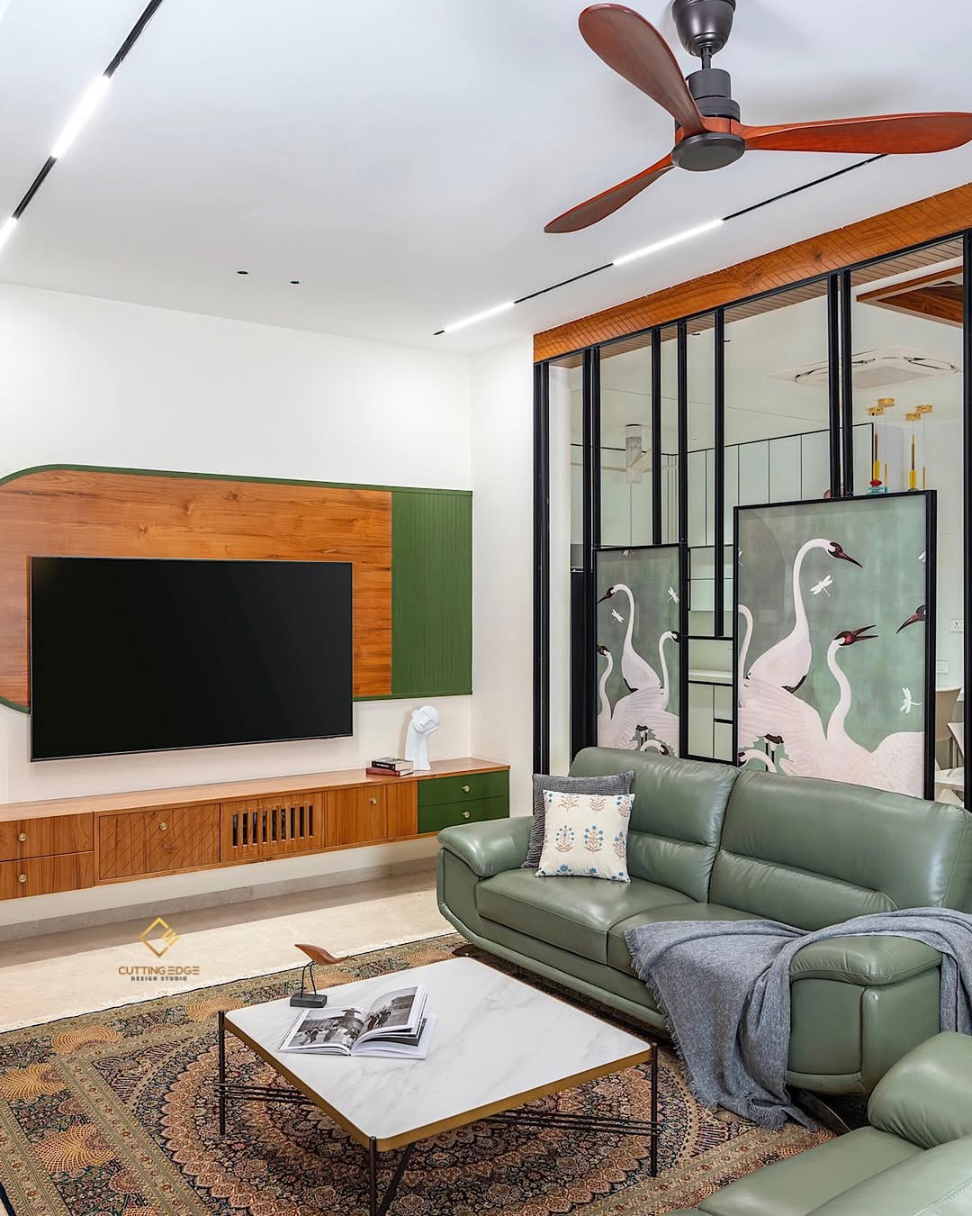 Modern living room with a green leather sofa, wall-mounted TV, wooden furniture, decorative crane-themed glass panels offering instant style and divider screen ideas, ceiling fan, and a coffee table with books on a patterned rug.