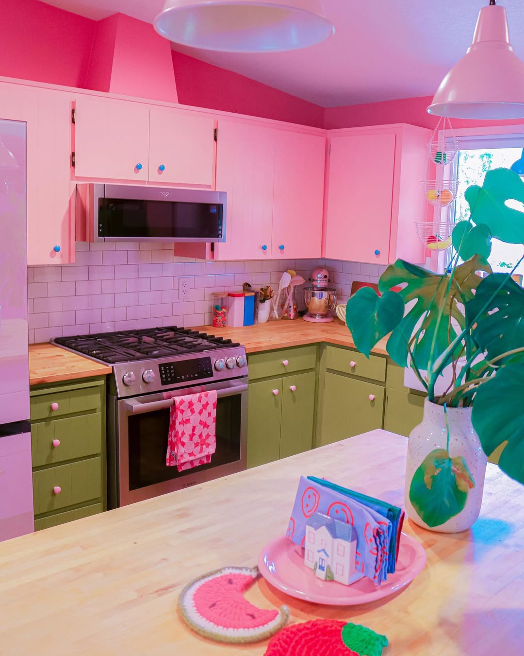 A pink kitchen with vibrant upper cabinets, green lower cabinets, stainless steel appliances, light wood countertops, and colorful decor items brightens the island and counters.