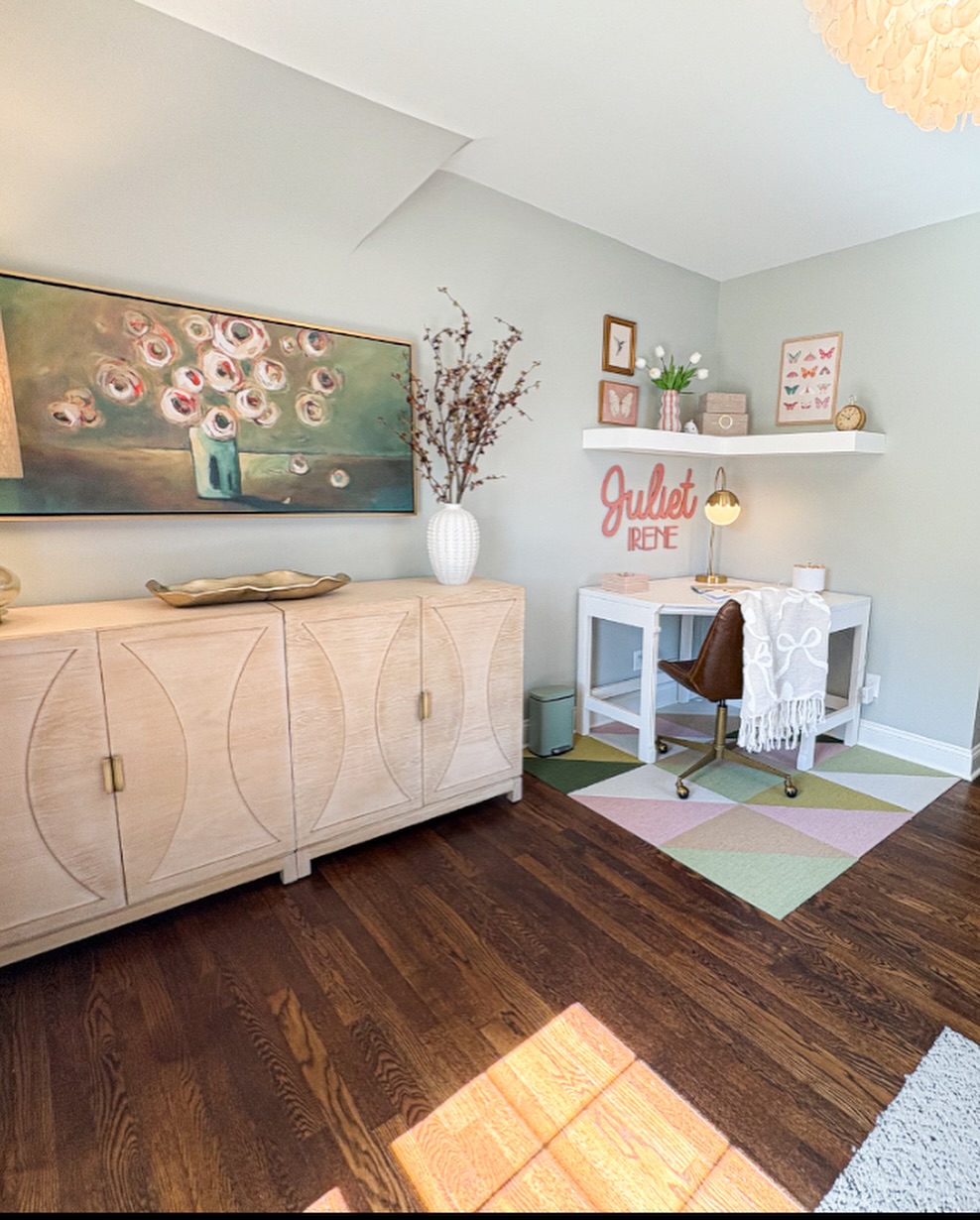 A small home office with a wooden cabinet, desk, brown chair, and shelves. Decor includes a flower painting, vases, a plant, and a sign reading