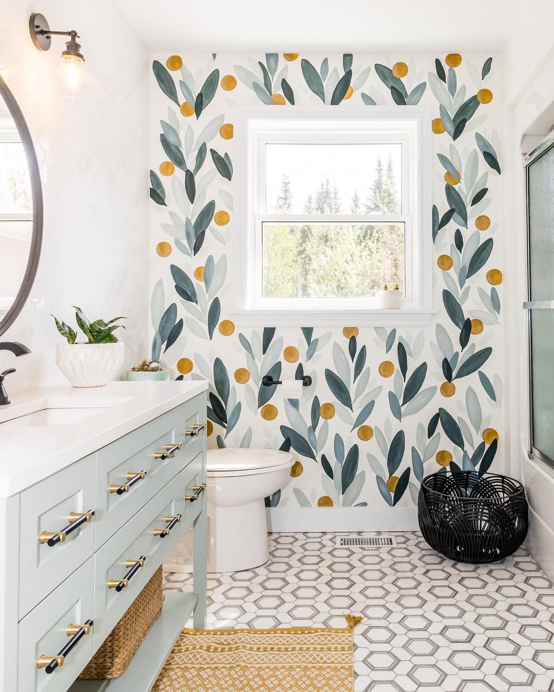 Bright small bathroom design featuring leaf and yellow dot patterned wallpaper, a mint green vanity, round mirror, hexagonal tile floor, and a window above the toilet—perfect inspiration for half bathroom ideas.