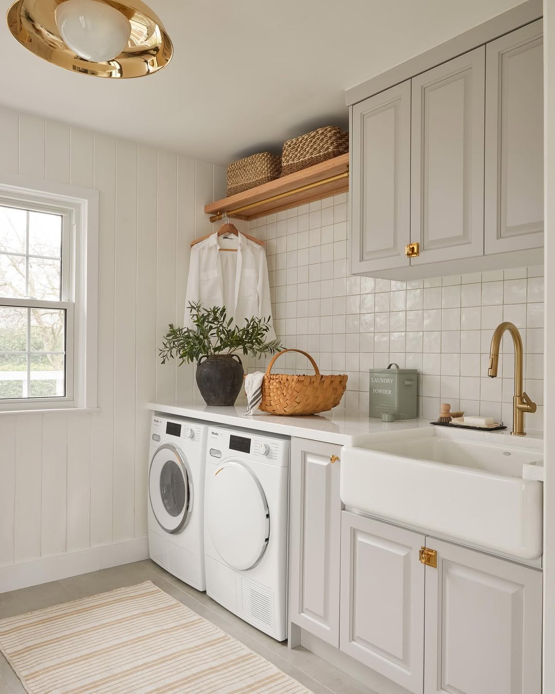 A cottage laundry room with a washer and dryer, a farmhouse sink, gray cabinets, a gold faucet, woven baskets, a potted plant, and a window letting in natural light.