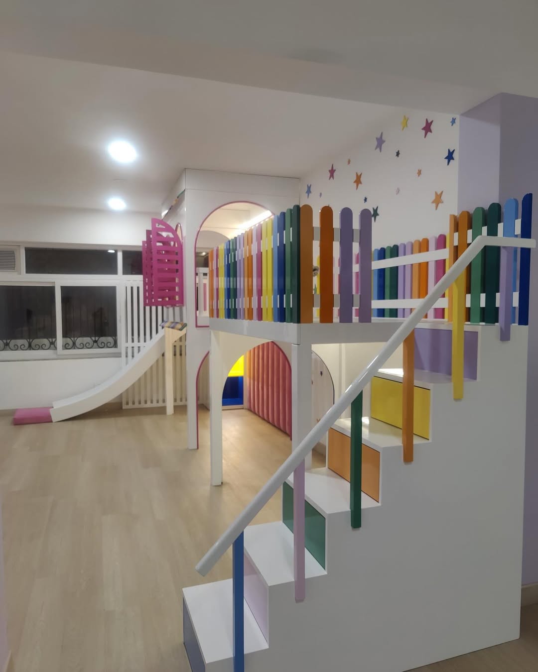Indoor play area with a colorful rainbow-themed staircase leading to an under stair playroom, slide, and fenced loft, set against light wood flooring and white walls with star decals.