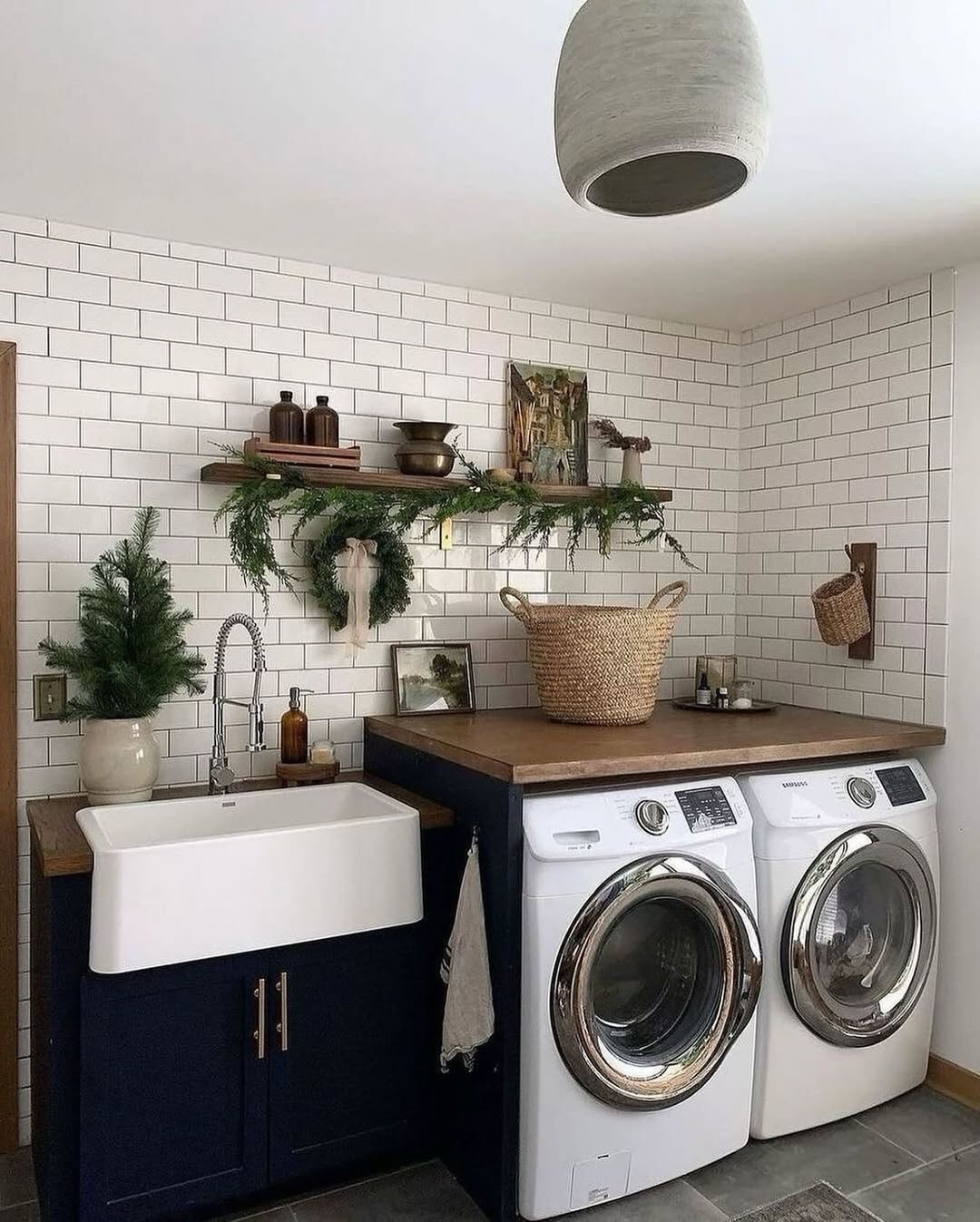 Cottage laundry room featuring a farmhouse sink, front-loading washer and dryer, wooden shelves, a wicker basket, and greenery decor against crisp white subway tile walls.