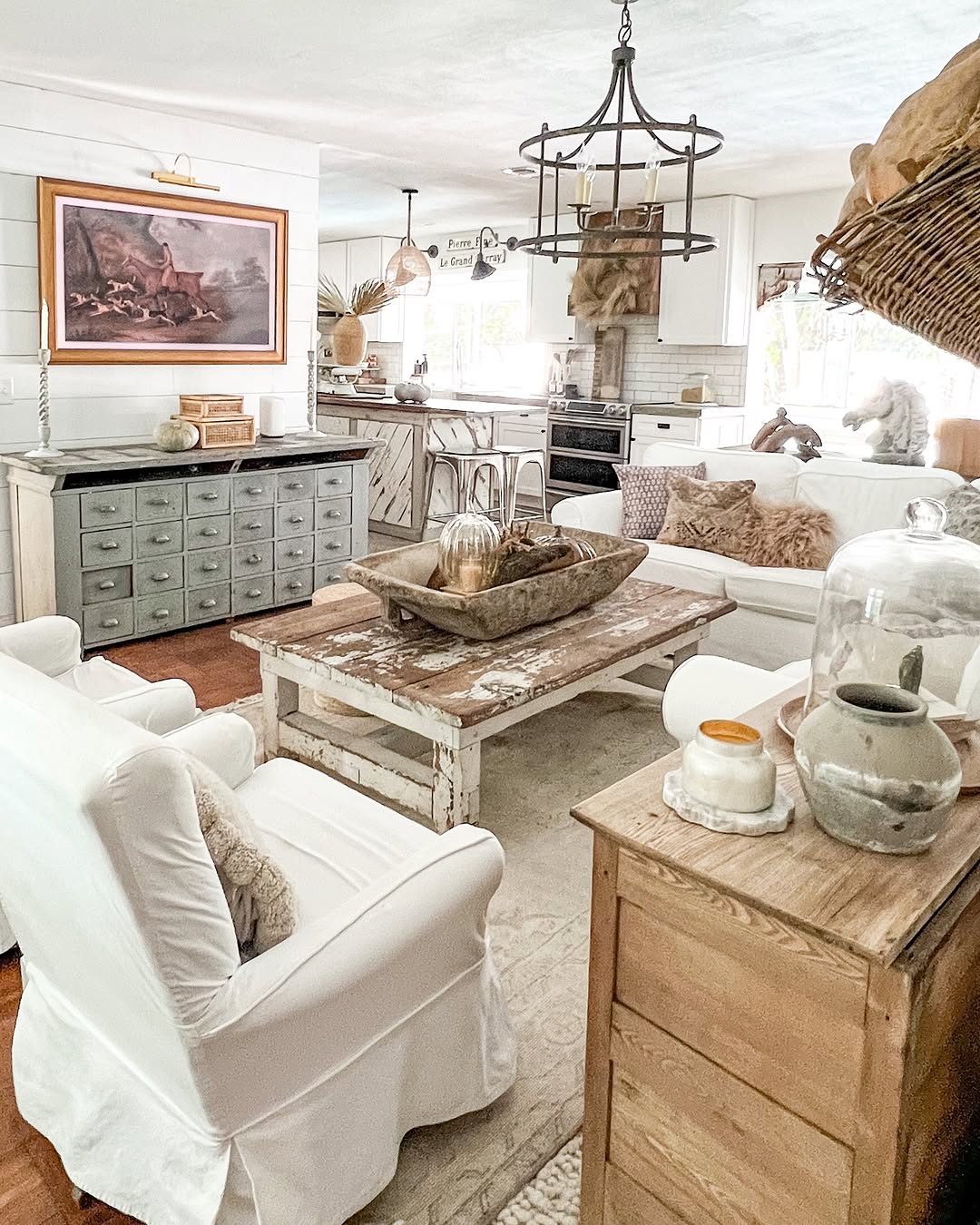 A rustic French country living room with white armchairs, a weathered wood coffee table, neutral decor, and a farmhouse-style kitchen in the background.