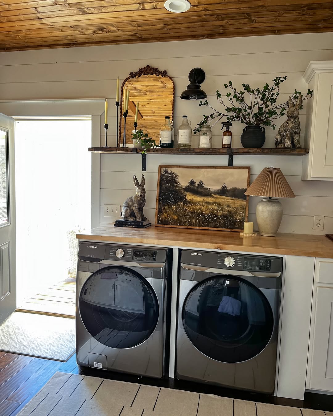 A cottage laundry room featuring two front-loading Samsung washers, wooden shelves adorned with decor, plants, and a framed landscape picture, all bathed in sunlight streaming through an open door.