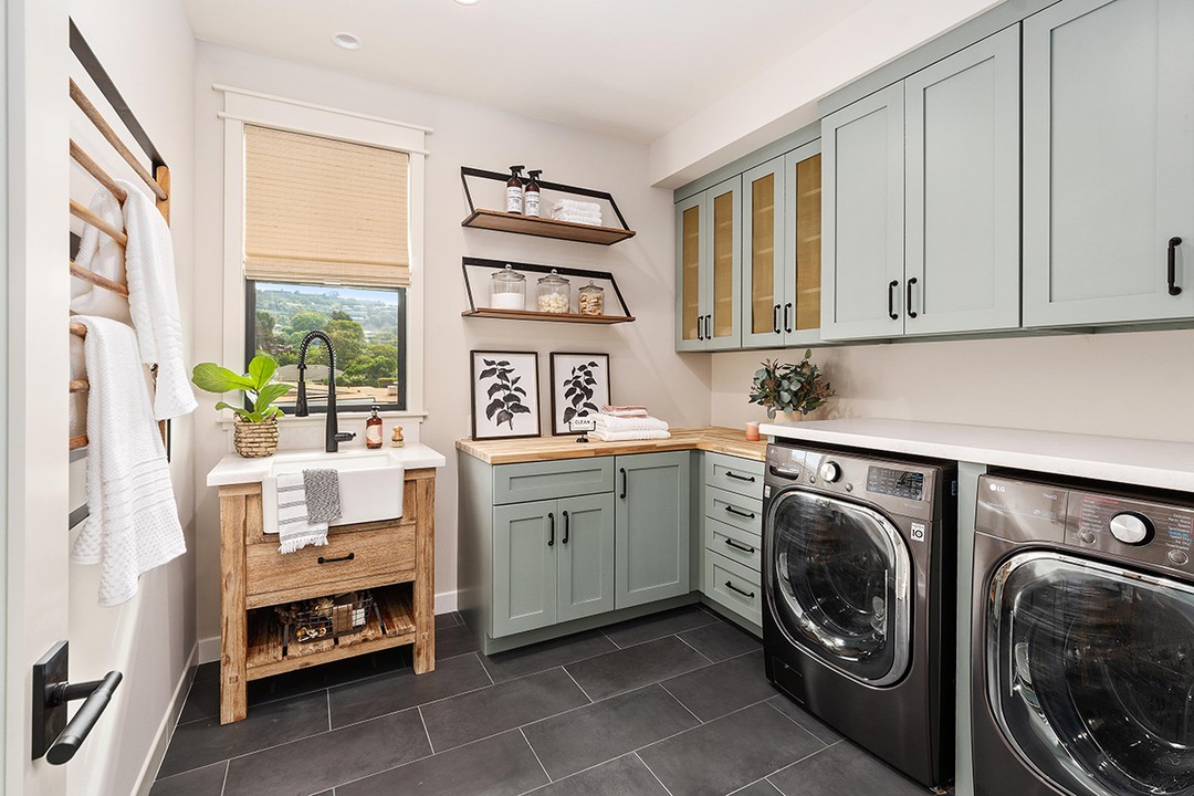 Modern cottage laundry room with light green cabinets, a farmhouse sink, wall shelves with jars, black washer and dryer, and a window letting in natural light.