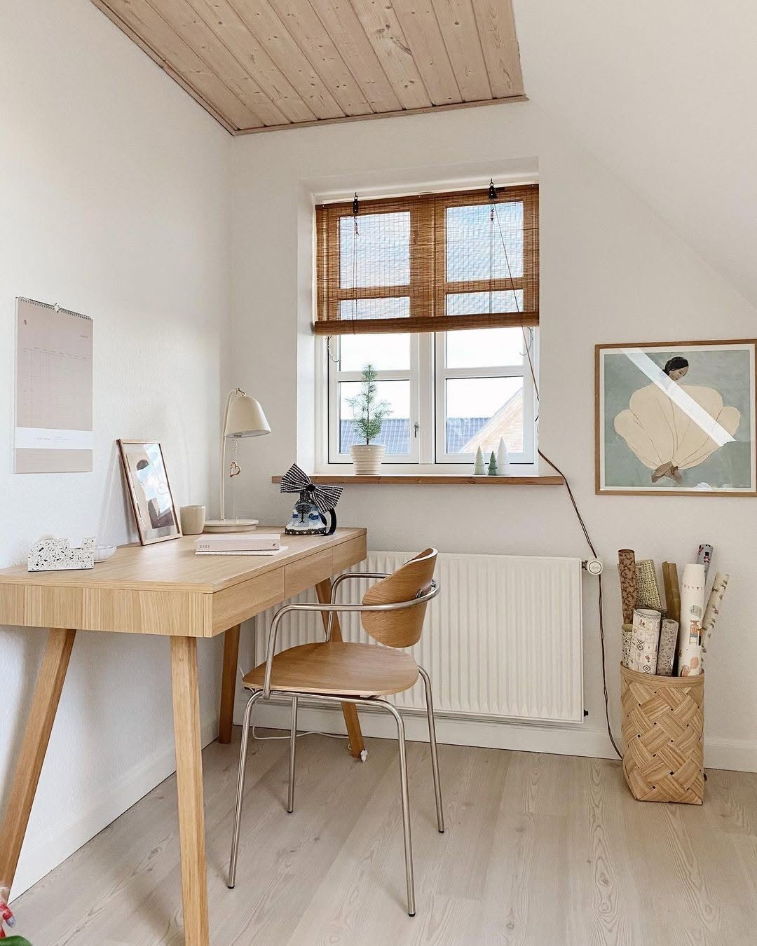 A minimalist small home office features a wooden desk, chair, lamp, calendar, framed art, and a basket of rolled paper by a window with blinds.