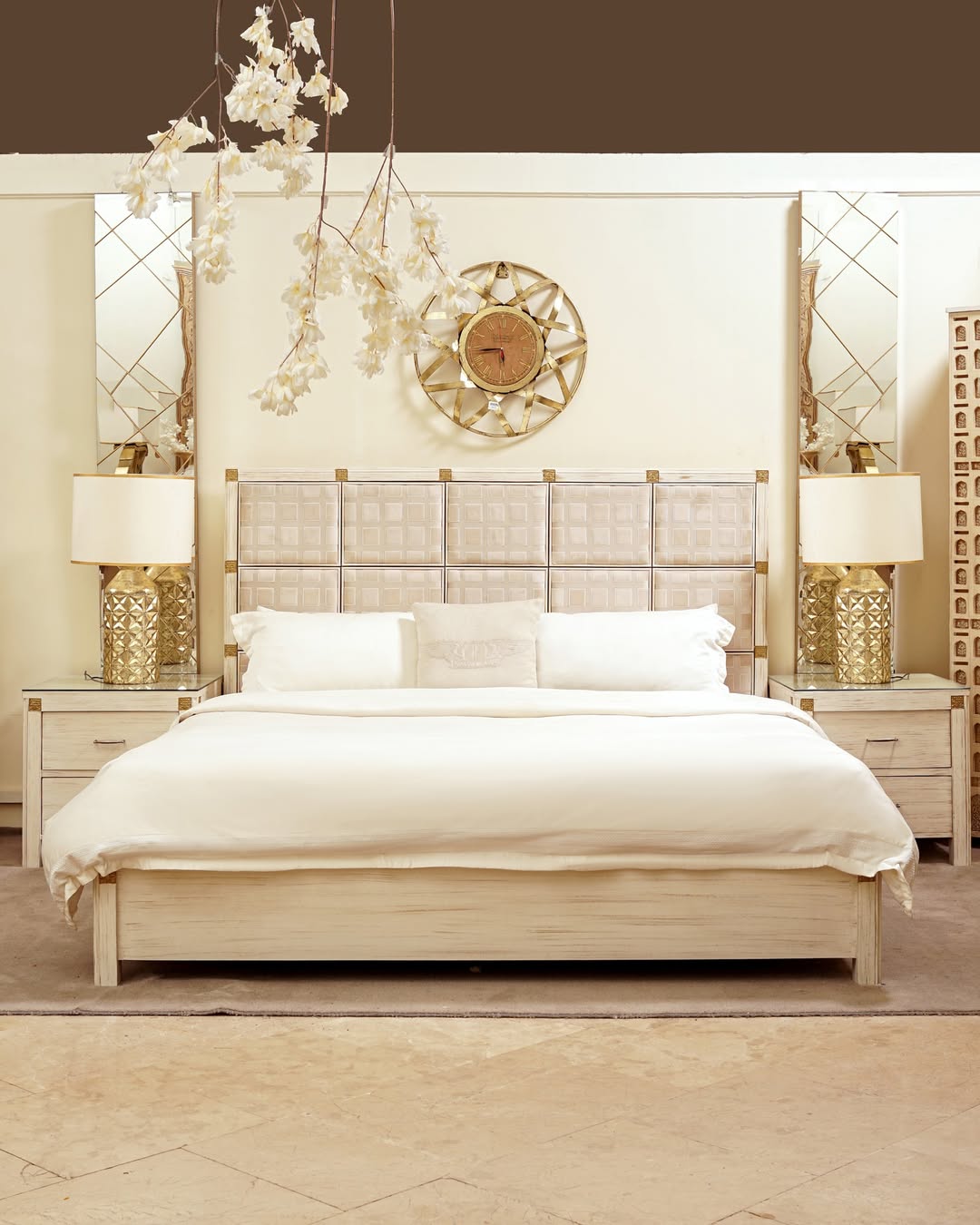 A modern bedroom with a large white bed featuring a stylish bedroom headboard, two mirrored nightstands with gold lamps, wall mirrors, a decorative wall clock, and hanging white flowers.