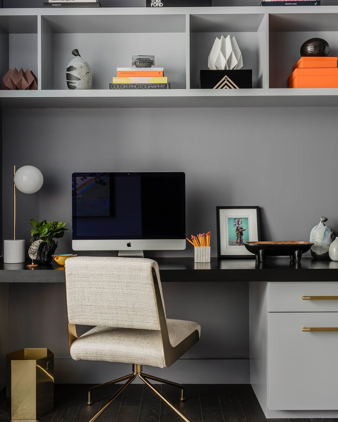 A modern small home office features a computer on a grey desk, a beige chair, shelves with books and decor, a lamp, and various desk accessories.