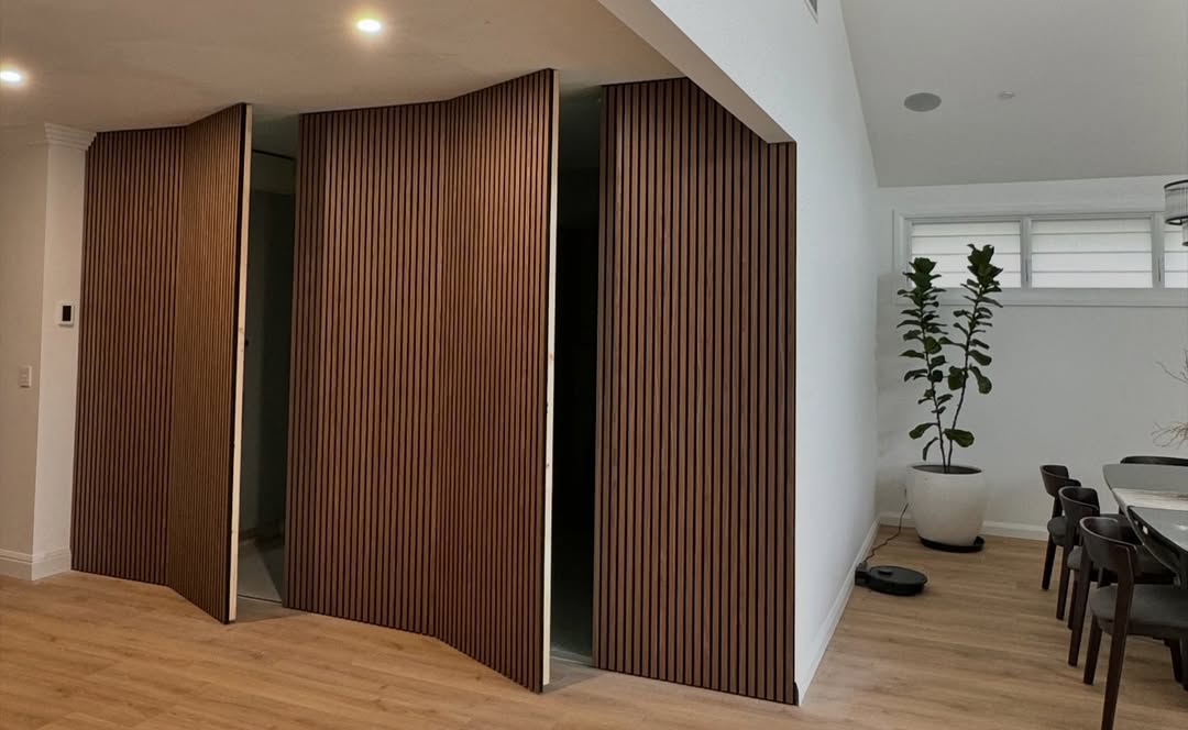 A modern interior with hidden doors crafted from folding wooden panels, light wood flooring, a potted plant, a dining table, and a robotic vacuum on the floor—perfect for stylish homeowners seeking hidden door ideas.
