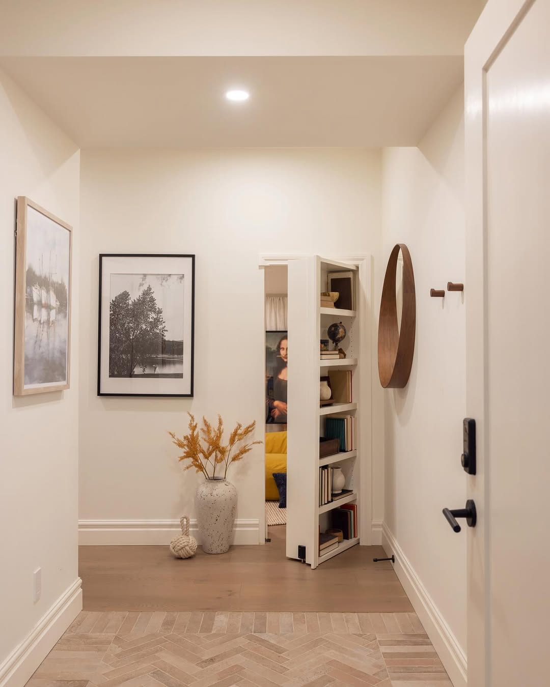 A home hallway features a partially open hidden door bookcase revealing a secret room; wall art, a vase with dried plants, and a round mirror add charm—perfect inspiration for stylish homeowners seeking unique hidden door ideas.